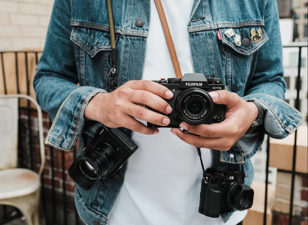 pexels-airam-datoon-12304035 - Expert Advice Person in denim jacket wearing multiple cameras around their neck and shoulders, with a Fujifilm X-T3 center and in their hands.