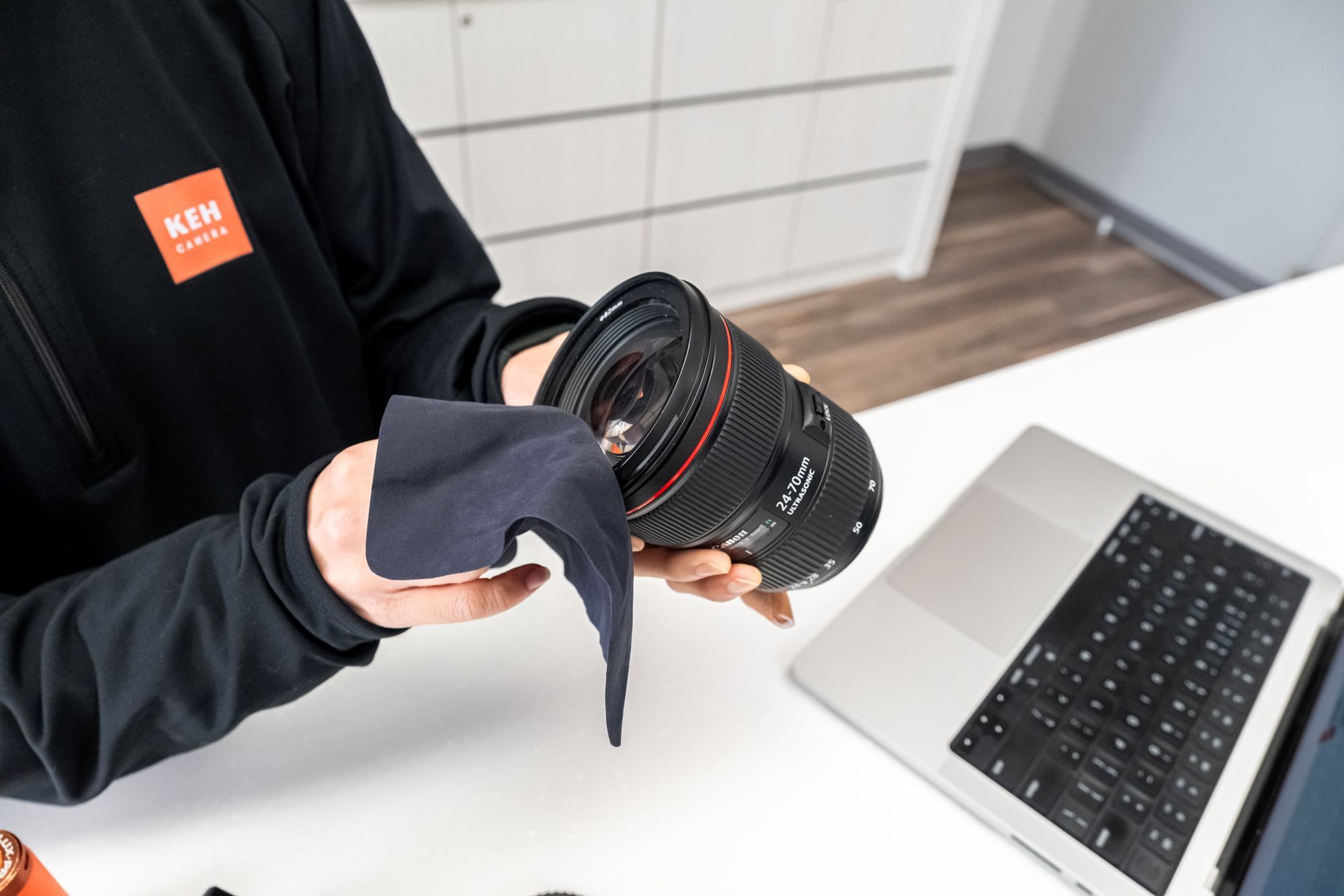 A KEH Camera team member in a black KEH jacket wipes a Canon 24-70mm lens with a microfiber cloth at a white counter, with a laptop nearby.