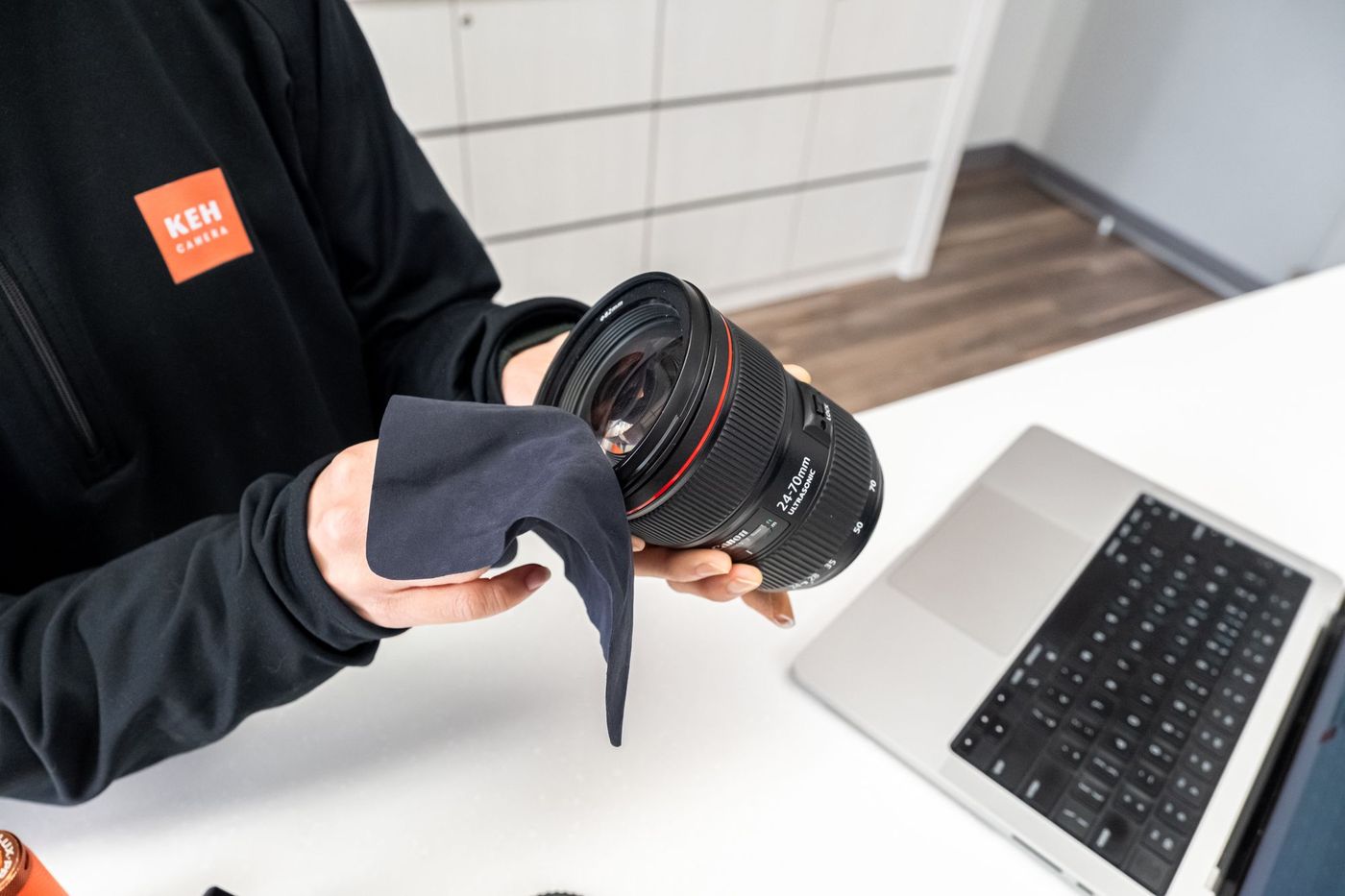 A KEH Camera team member in a black KEH jacket wipes a Canon 24-70mm lens with a microfiber cloth at a white counter, with a laptop nearby.