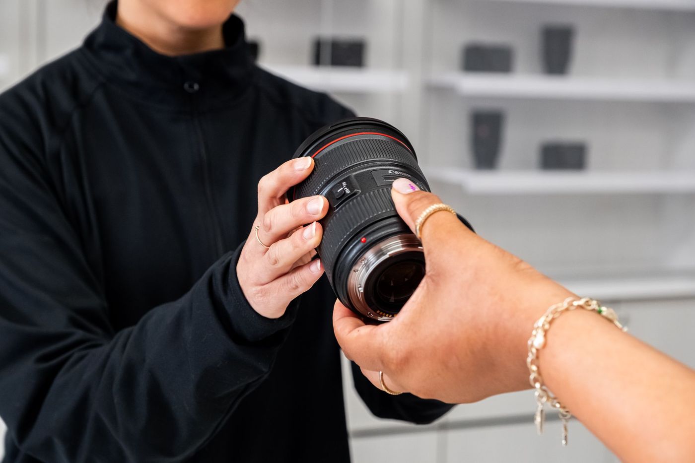 Close-up of a KEH Camera team member and a customer's hands holding a Canon 24-70mm lens, with the rear lens mount and contacts visible.